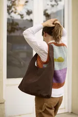 Cartera tipo tote de tamaño mediano, confeccionada en cuero suave color marrón oscuro. Presenta un diseño minimalista con costura central vertical y un pañuelo estampado anudado en una de las asas.