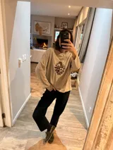 Sweater de punto color beige con cuello redondo y diseño de escudo bordado en el frente. Presenta un corte holgado y mangas largas.