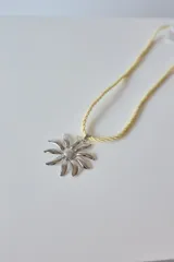 Collar con tiento trenzado color crema y dije de acero quirúrgico hipoalergénico con forma de sol o flor estilizada.