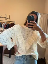 Camisa de encaje color crudo con cuello alto y volados frontales que recorren el pecho y los hombros. Presenta mangas largas con puños acampanados y un diseño romántico y delicado.