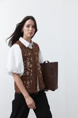 Chaleco tejido color marrón con bordados artesanales de motivos florales y geométricos en color crema. Presenta cuello redondo, cierre frontal con botones y diseño sin mangas.