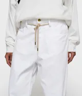 Pantalón blanco de corte recto y holgado, confeccionado en algodón orgánico con certificación GOTS. Presenta cintura ajustable con cordón beige, cierre de botón y cremallera, y bajos sin rematar con efecto deshilachado.