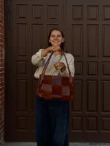 Cartera de hombro confeccionada en cuero y gamuza con diseño de damero, combinando paneles de texturas lisas y aterciopeladas. Presenta una manija trenzada y detalles de costuras artesanales en los laterales.