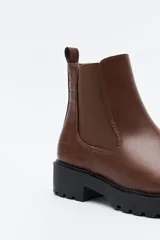 Bota tipo chelsea de caña baja con suela dentada robusta. Presenta un diseño combinado con paneles laterales elásticos, acabado liso en el frente y detalle texturizado en relieve en la parte posterior, incluyendo un tirador trasero para facilitar el calce.