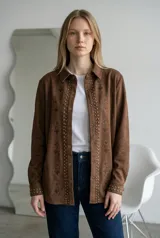 Sobrecamisa de gamuza color marrón chocolate, con cierre frontal de botones metálicos, detalle de tachas doradas a lo largo de la botonadura y puños, y bordados florales en los paneles frontales.