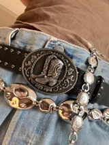 Cinturón negro de cuero sintético con hebilla plateada con diseño de botas cowboy y tachas plateadas.