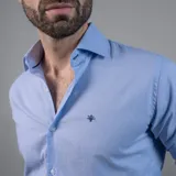 Camisa de vestir celeste de manga larga, con cuello clásico y botones blancos. Presenta un logo bordado en azul oscuro en el pecho.