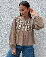 Camisa de color marrón con panel frontal bordado con motivos florales y geométricos. Presenta cuello redondo, cierre frontal con botones forrados, mangas largas con puños elásticos y un diseño con frunces bajo el canesú.
