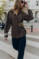 Camisa de cuello solapa y manga larga, color marrón con rayas verticales blancas. Presenta cierre frontal con botones ocultos y dos hebillas decorativas en color beige en el lateral.