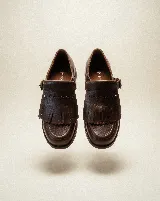 Mocasín de cuero marrón con diseño artesanal, que presenta una solapa desmontable con flecos y una hebilla lateral metálica. Su diseño permite transformar el calzado, ofreciendo versatilidad entre un estilo con flecos y uno más minimalista.