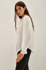 Camisa blanca de cuello alto con volados y encaje, mangas largas abullonadas con puños de encaje. Presenta detalles de encaje y pliegues verticales en el frente.