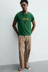 Camiseta de corte relajado, color verde, con cuello redondo y manga corta. Presenta un texto bordado en relieve con letras amarillas y contorno oscuro en el centro del pecho.