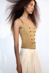 Top corsetero de color tostado con escote cuadrado, tirantes anchos y cierre de cremallera en la espalda. Cuenta con costuras marcadas y filas de botones metálicos dorados decorativos en el frente.
