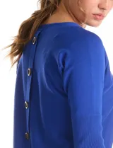 Buzo de tejido fino con cuello redondo, manga tres cuartos con puños acanalados y detalle de pespunte vertical en el frente. Presenta un diseño liso con aplique de botones en la espalda.