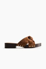 Sandalias estilo zueco color marrón, con tiras de gamuza anudadas en la parte delantera y detalles de tachas metálicas doradas en los laterales. Cuenta con suela de madera oscura de tacón cuadrado bajo.