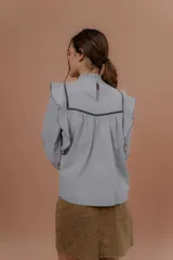Blusa de jeans fino color celeste con detalles de terciopelo negro en contraste. Presenta cuello alto con volados, mangas largas abullonadas con puños ajustados y volados decorativos en los hombros y el frente.