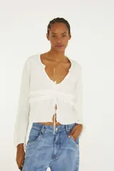 Blusa blanca de manga larga con escote en V, detalles de volados horizontales y cierre frontal con tiras ajustables.