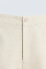 Bermuda de corte relajado color beige claro, confeccionada en tejido con textura. Presenta cierre frontal con cremallera y botón, bolsillos delanteros y bolsillos traseros de doble vivo con botón.