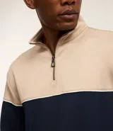 Buzo de media malla con diseño de bloques de color, presentando la parte superior en beige y la inferior en azul marino, separadas por una línea blanca. Cuenta con cuello alto con cierre frontal parcial y puños acanalados.