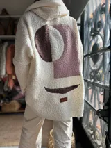 Campera con capucha de textura tipo corderito en color manteca, con diseño abstracto en la espalda compuesto por formas geométricas en tonos marrón y rosado. Presenta un corte holgado y una etiqueta de marca en la parte inferior.