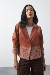 Blazer de gamuza color terracota con bordados decorativos en color crudo en solapas, puños y ruedo. Presenta corte recto, solapas clásicas y cierre frontal con un botón forrado.