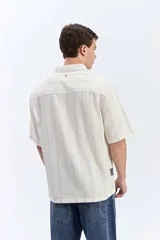 Camisa de manga corta oversize en tejido rústico, con diseño de rayas verticales celestes y blancas, cuello clásico y cierre frontal de botones.