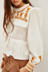 Camisa de algodón celeste con detalles de volados en los hombros y bordados en color negro en el pecho y las mangas. Presenta cierre frontal con botones y puños ajustados.