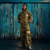 Pantalón de jogging Corteiz con estampado de camuflaje digital en tonos verdes y marrones. Presenta cintura elástica y un logo gráfico ovalado estampado en color blanco en la pierna izquierda.