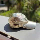 Gorra de cuero metalizado dorado, con ojales metálicos decorativos y etiqueta de marca aplicada en el lateral.