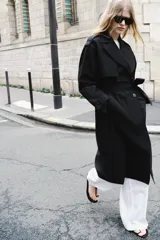 Trench largo de color negro con cuello de solapa, manga larga con trabillas ajustables y cierre frontal cruzado con doble botonadura. Cuenta con bolsillos delanteros de vivo, solapa cortavientos en el pecho y cinturón ajustable del mismo tejido.