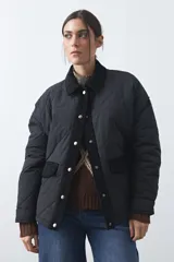 Campera tipo barn capitoneada de color marrón, con silueta corta y relajada. Presenta cuello de pana, cierre frontal con broches metálicos y bolsillos laterales funcionales.
