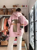 Campera con capucha confeccionada en tejido tipo corderito color rosa, con diseño de patchwork en la espalda que combina parches de tela lisa, texturizada y estampada en tonos verde oliva, marrón y rosa. Presenta un corte holgado y etiqueta de marca en el ruedo.