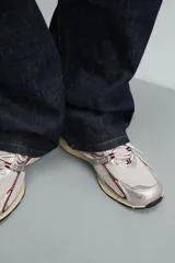 Zapatilla deportiva tipo running con diseño técnico, confeccionada en una combinación de materiales sintéticos y malla. Presenta una estética metalizada en tonos plata con detalles en color granate y una suela con volumen de diseño irregular en tono crema.