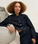 Camisa de jean estilo cropped de color azul oscuro, con cuello inglés, mangas largas, cierre frontal de botones metálicos y un bolsillo aplicado en el pecho. Cuenta con costuras pespunteadas en tono contrastante.