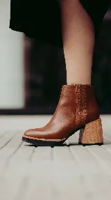 Bota corta de cuero color marrón con acabado liso, que presenta un detalle de textura acanalada en el lateral y cierre interno. Cuenta con un taco ancho y estable de color negro.