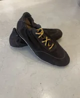 Zapatilla tipo 'champion' de gamuza color marrón oscuro, diseño minimalista y liviano, con cordones bicolores (amarillo y negro). Presenta un taco oculto y construcción en cuero.