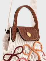 Bolso de mano tipo cesta de la línea Le Pliage Mesh de Longchamp, en tamaño XS. Presenta una estructura de red tejida en color marfil con detalles de lazos bordados en tonos pastel y asas cortas de cuero marrón. Incluye una correa larga ajustable para llevar al hombro o cruzado.