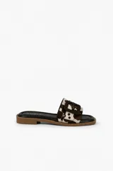 Sandalia plana marca Daniel Cassin con estampa animal print tipo vaca de efecto pelo, capellada ancha con hebilla grande revestida y suela baja con base marrón.