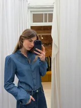 Camisa de denim fino azul, con cuello alto con volado, mangas largas abullonadas en los hombros, puños con volado y cierre frontal con botones a contratono en color rojo.