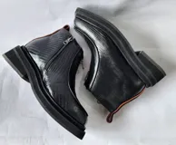 Bota corta de cuero negro con diseño de punta cuadrada y suela track. Presenta un cierre frontal central, detalles de pespuntes y una textura combinada en los laterales.