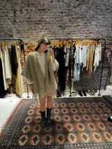 Ruana tejida a mano en lana merino color beige, con textura de punto y detalle de flecos en el ruedo. Presenta sutiles destellos brillantes integrados en el tejido.