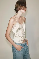 Top crop satinado de color crudo, con escote en pico, tirantes finos y detalle de encaje combinado en el escote y al frente. Presenta cierre frontal con pequeños botones y bajo de corte asimétrico.