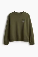 Buzo oversized color verde militar con interior cepillado suave. Presenta hombros caídos y un diseño de texto bordado en el pecho que dice "THE LAKE DISTRICT" con un pato. Tiene ribete acanalado en cuello, puños y bajo.
