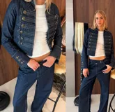 Chaqueta de denim estilo militar con diseño napoleónico, caracterizada por su doble fila de botones metálicos decorativos en el frente y mangas acampanadas con detalles de botones en los puños. Presenta un corte entallado y cuello redondo.