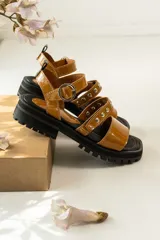 Sandalias de cuero color suela con acabado charolado, diseño de tiras con tachas metálicas y hebilla ajustable. Poseen una suela de goma negra tipo track con plataforma.