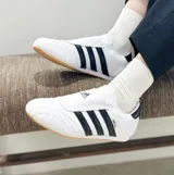 Championes Adidas modelo Taekwondo, color blanco con las icónicas tres tiras laterales en negro. Presentan un diseño minimalista sin cordones, con una lengüeta que cubre el empeine y suela de goma color caramelo.