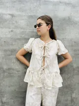 Blusa estampada con hojas verdes y blancas, con escote en V y mangas cortas abullonadas. Presenta volados en el ruedo y detalle de lazos en el frente.