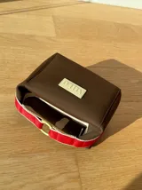 Neceser de neopreno color borgoña, con cierre superior y forro interior. Tiene un asa de mano con una franja azul y blanca.