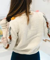 Sweater tejido a mano en punto jersey de 100% lana merino, color blanco con detalles texturizados en punto bucle de tonos rosados, mostaza, marrón y gris, formando líneas onduladas en el frente y las mangas. Cuenta con cuello redondo base y puños largos acANALados.