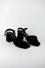 Sandalia de tacón cuadrado medio, confeccionada en material textil tipo gamuza color negro. Presenta un diseño de tiras cruzadas con nudo en la parte delantera y una tira ajustable con hebilla metálica en el tobillo.